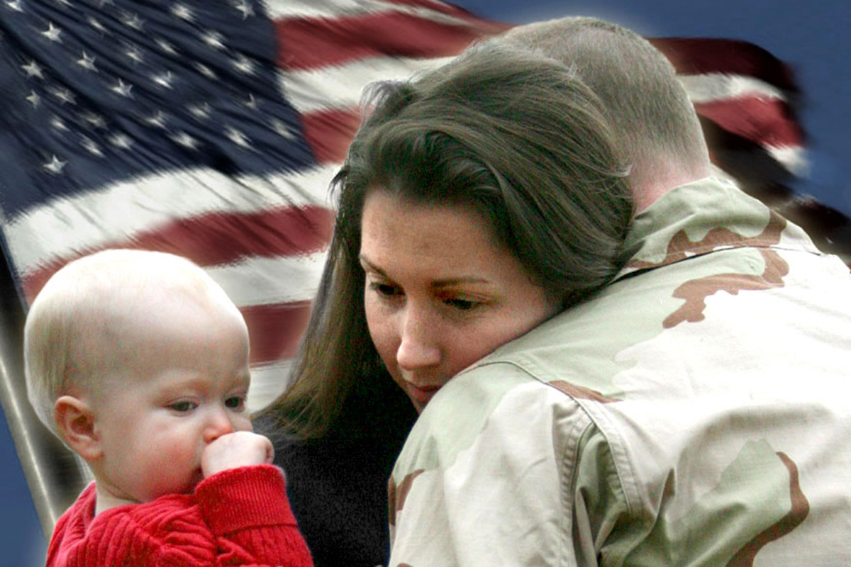 Serviceman hugging his wife and baby while Old Glory waves in the background