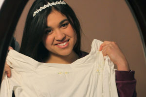 Dark haired dark eyed tan skinned young woman wearing sparkling headband holding up her wedding dress before a mirror preparing to be a help meet
